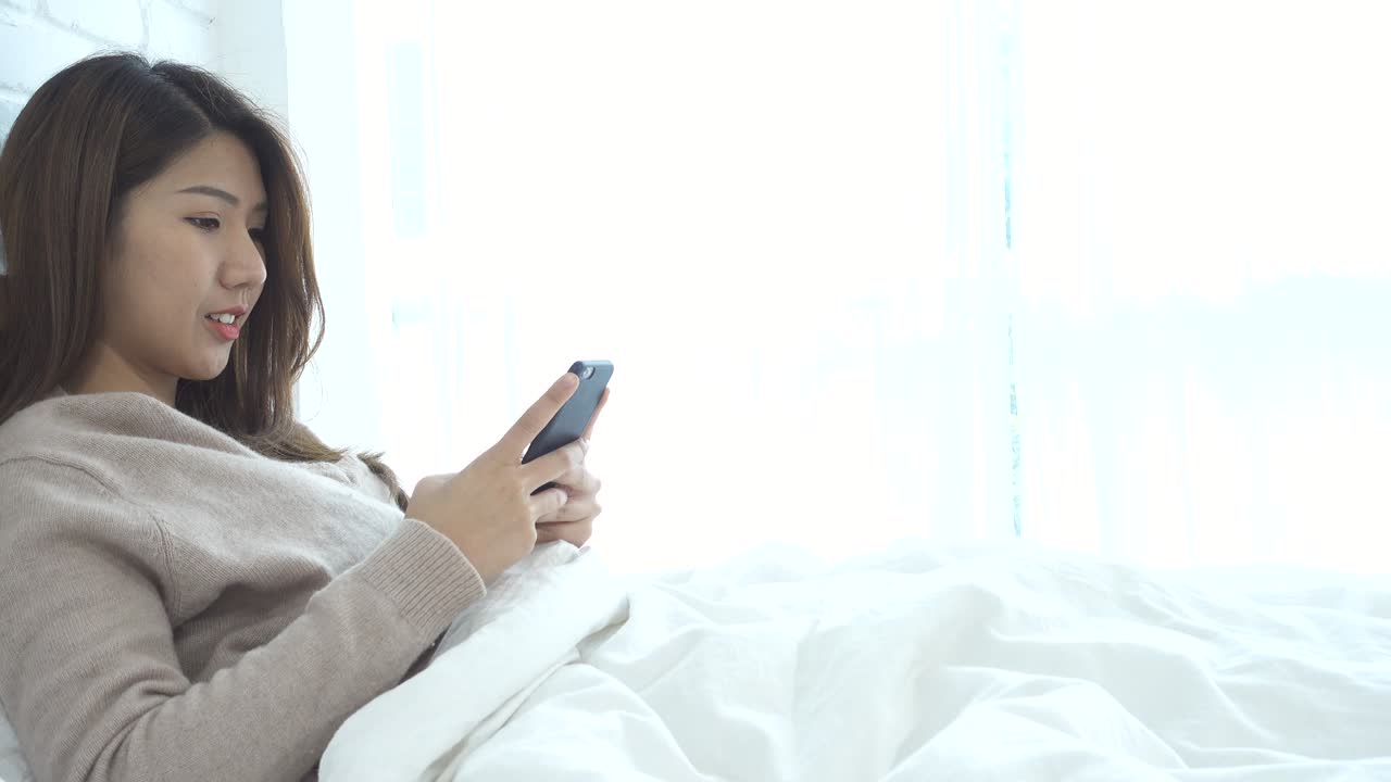 행복한 아시아 여성은 아침에 침대에서 스마트 폰을 사용합니다. 아시아 여성은 침대에서 스마트폰으로 소셜 앱을 확인합니다. 웃는 여성은 집에서 드폰으로 인터넷을 서핑합니다. 모바일 중독자 개념.