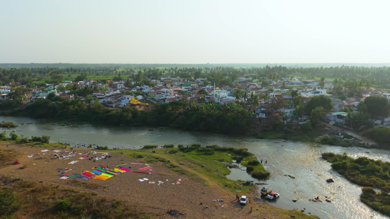 Vista aerea di un villaggio rurale lungo il fiume che mostra persone che lavano i vestiti, veicoli puliti vicino alla riva, acqua che scorre calma, campi verdi, case del villaggio e atmosfera di campagna tranquilla
