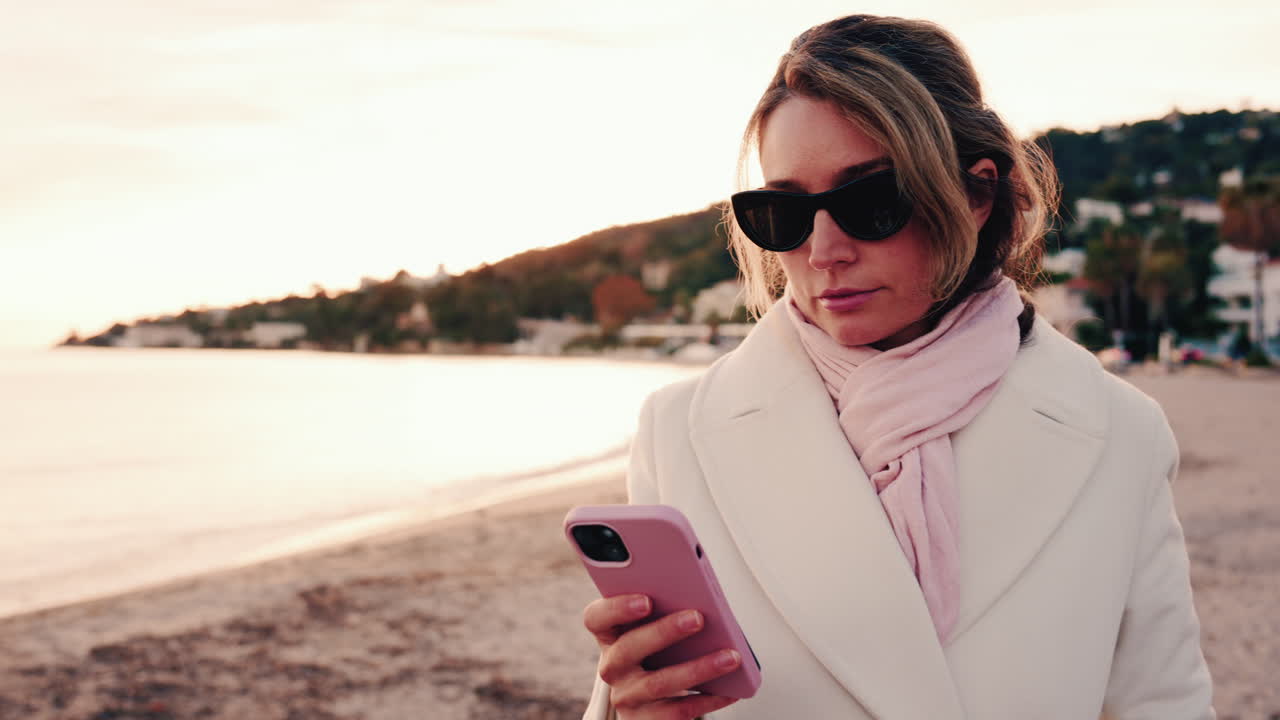 Donna elegante con cappotto bianco, sciarpa e occhiali da sole che controlla il telefono mentre è in piedi su una spiaggia al tramonto