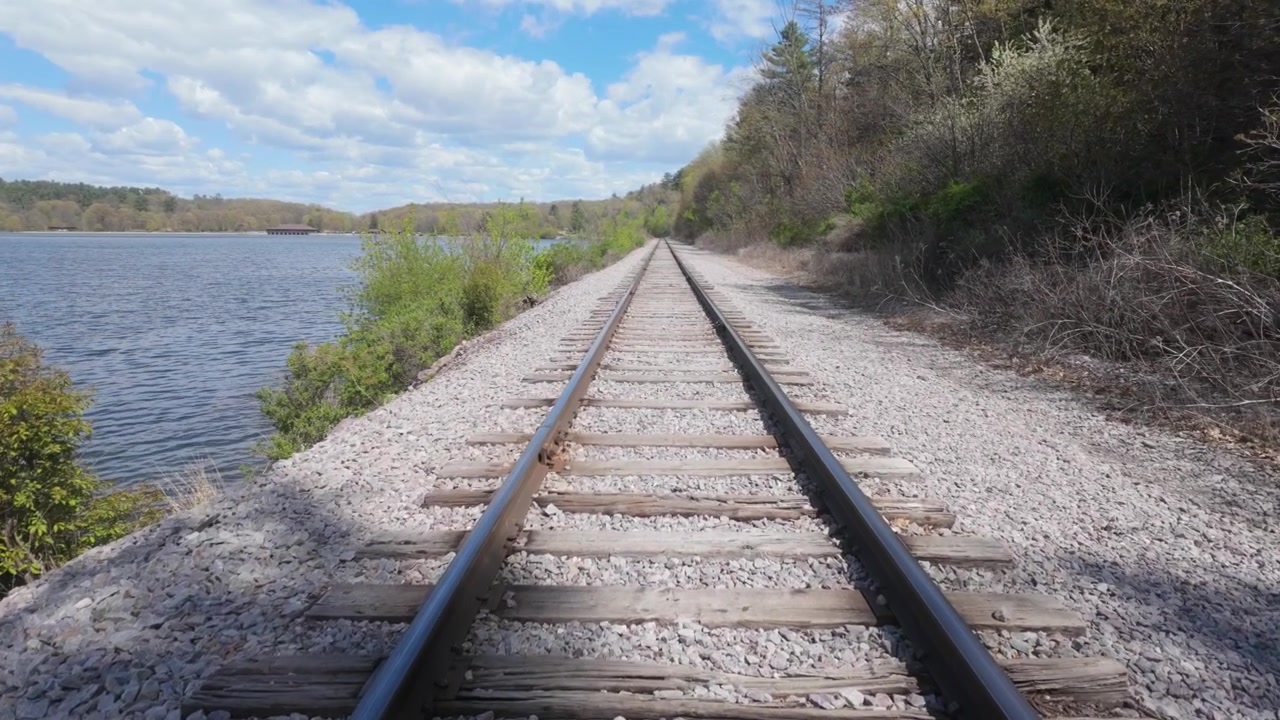 Caminhando pelos trilhos do trem em vista em primeira pessoa. Os trilhos são cercados por água de um lado e árvores e folhagem do outro lado em um lindo dia na natureza