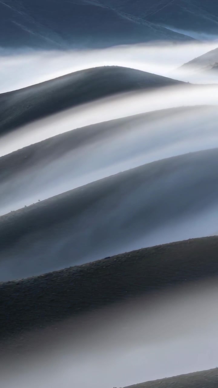 Vertical video: Shifting fog drifting over layered ridges in mountain valley, driven by light wind