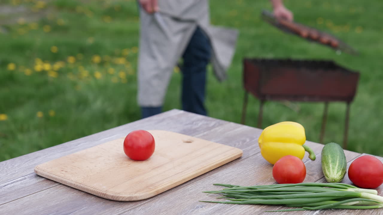 Fresh vegetable on the table in the open air. Tomatoes, cucumbers, onion and pepper on table on the background of a man cooking sausages on grill.