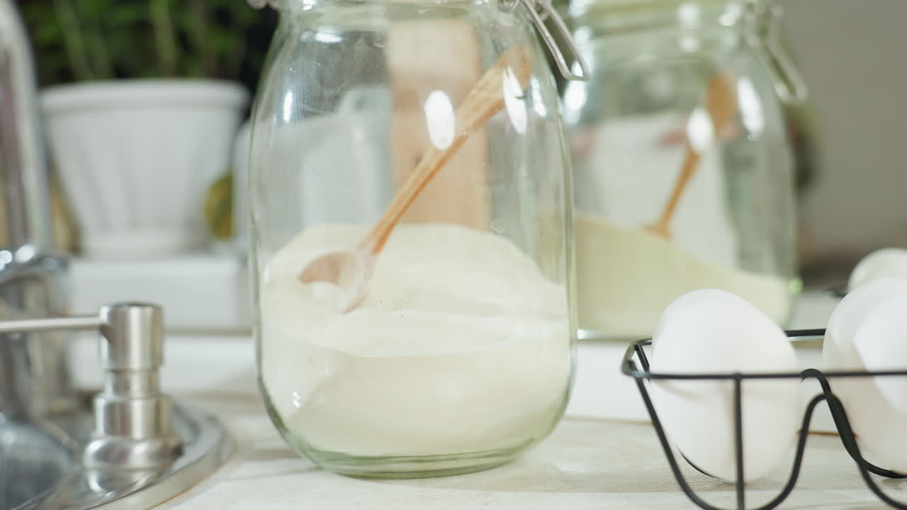 Glass jar filled with white flour and wooden spoon placed on tidy kitchen counter near eggs and sink, with soft daylight reflecting surroundings, while someone walks in and drops white cup