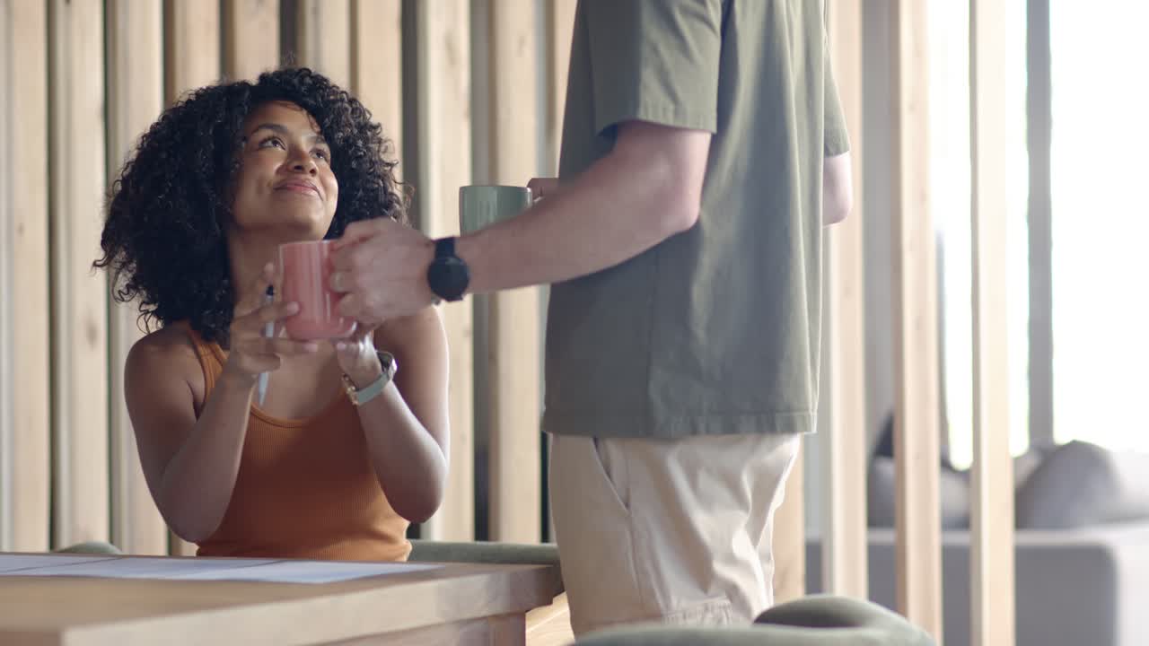 Couple diversifié dans la cuisine, l'homme entrant avec des tasses, offrant son aide à la femme qui prend une tasse en souriant, avec un espace de copie