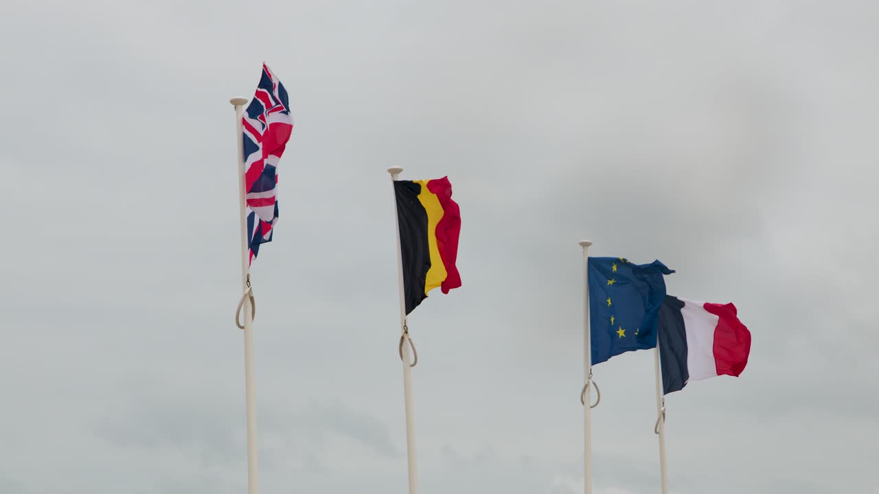Four national flags wave on tall poles in a windy coastal area beneath cloudy skies