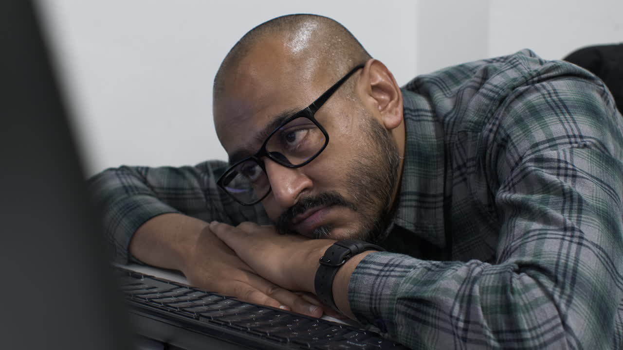 Close up shot of an exhausted young Indian entrepreneur resting his head on his arms in front of a computer, eyes fixed on the screen waiting for confirmation that the business proposal has been sent