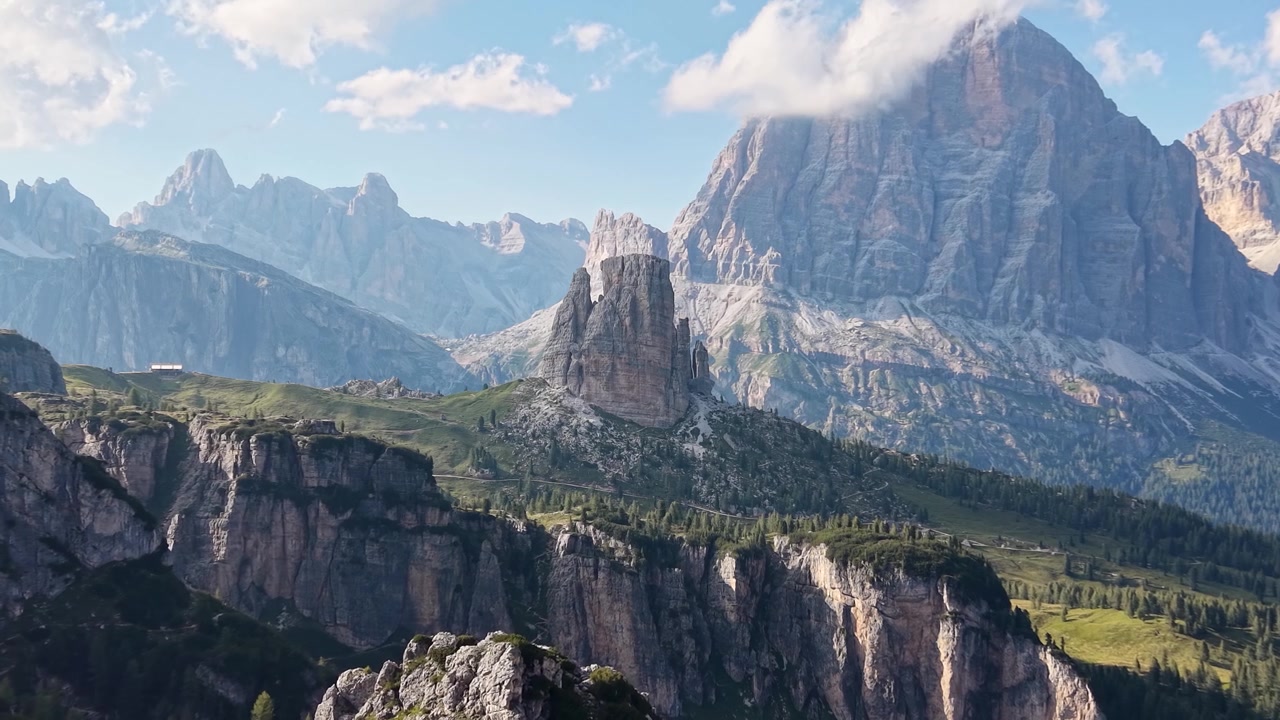 Veduta aerea con drone delle caratteristiche formazioni rocciose delle Cinque Torri vicino al Passo Giau, circondate da aspre cime calcaree, prati alpini e pendii boscosi sotto un cielo parzialmente nuvoloso