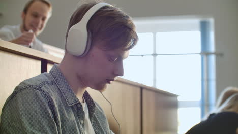 a student sitting in a university auditorium with a large group of people sits in headphones and listens to music and looks at the smartphone screen. it is in a state of calm. class break