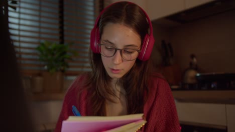 A confident brunette girl in red wireless headphones and glasses sits in the kitchen in a modern apartment and writes down an online lesson in her large notebook
