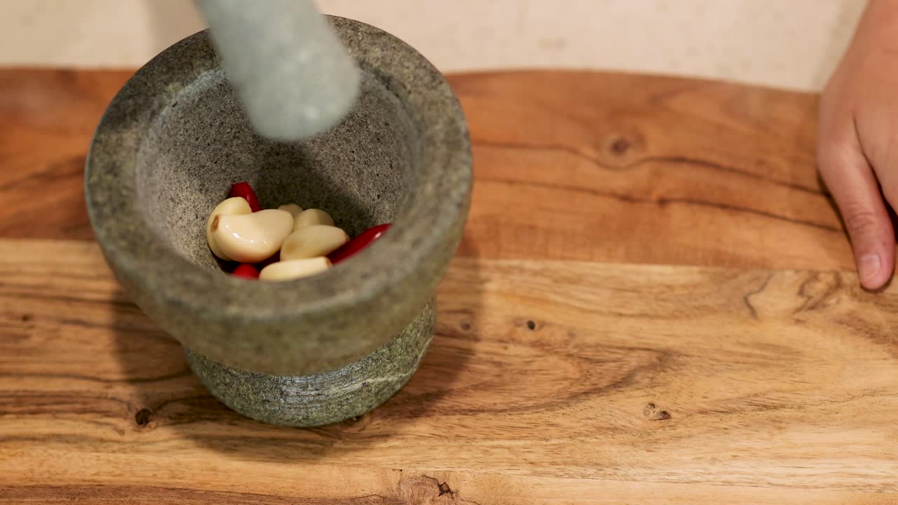 Hands pound garlic and chili in a stone mortar on a wooden surface, showcasing traditional culinary preparation