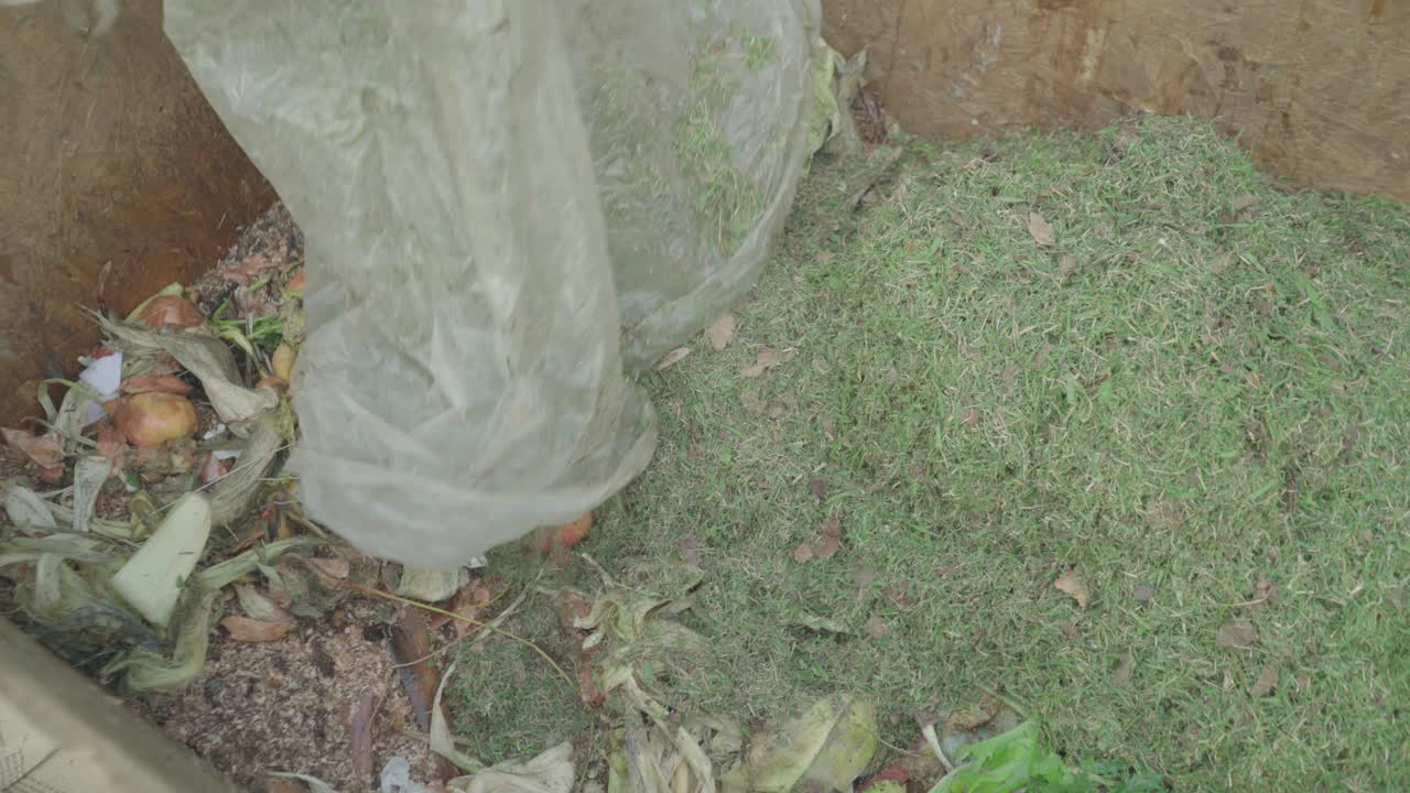 Women prepare a home wooden compost heap box. They add grass and scatter it around the box.