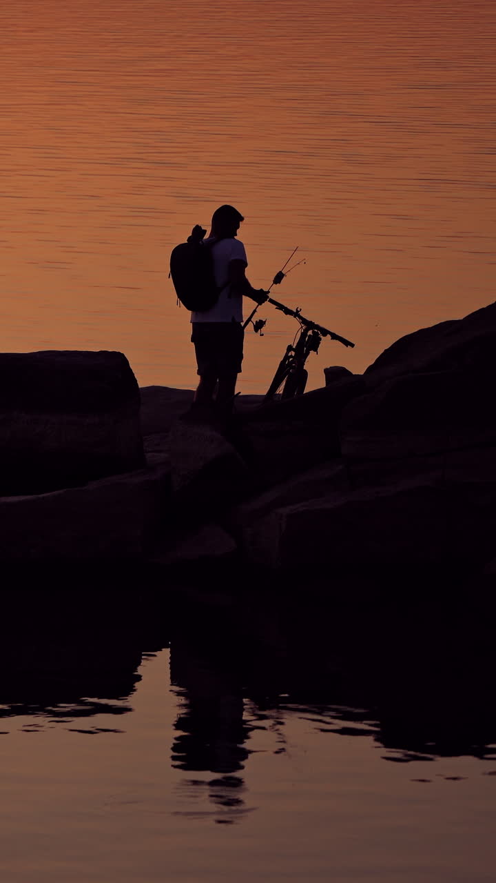 Man with fishing rods near his bike on the evening water background. Silhouette of a fisherman with rucksack is gathering home standing on stony rocks over water. Vertical video