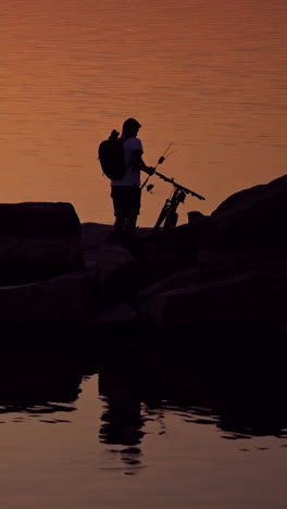 man with fishing rods near his bike on the evening water background. silhouette of a fisherman with rucksack is gathering home standing on stony rocks over water. vertical video