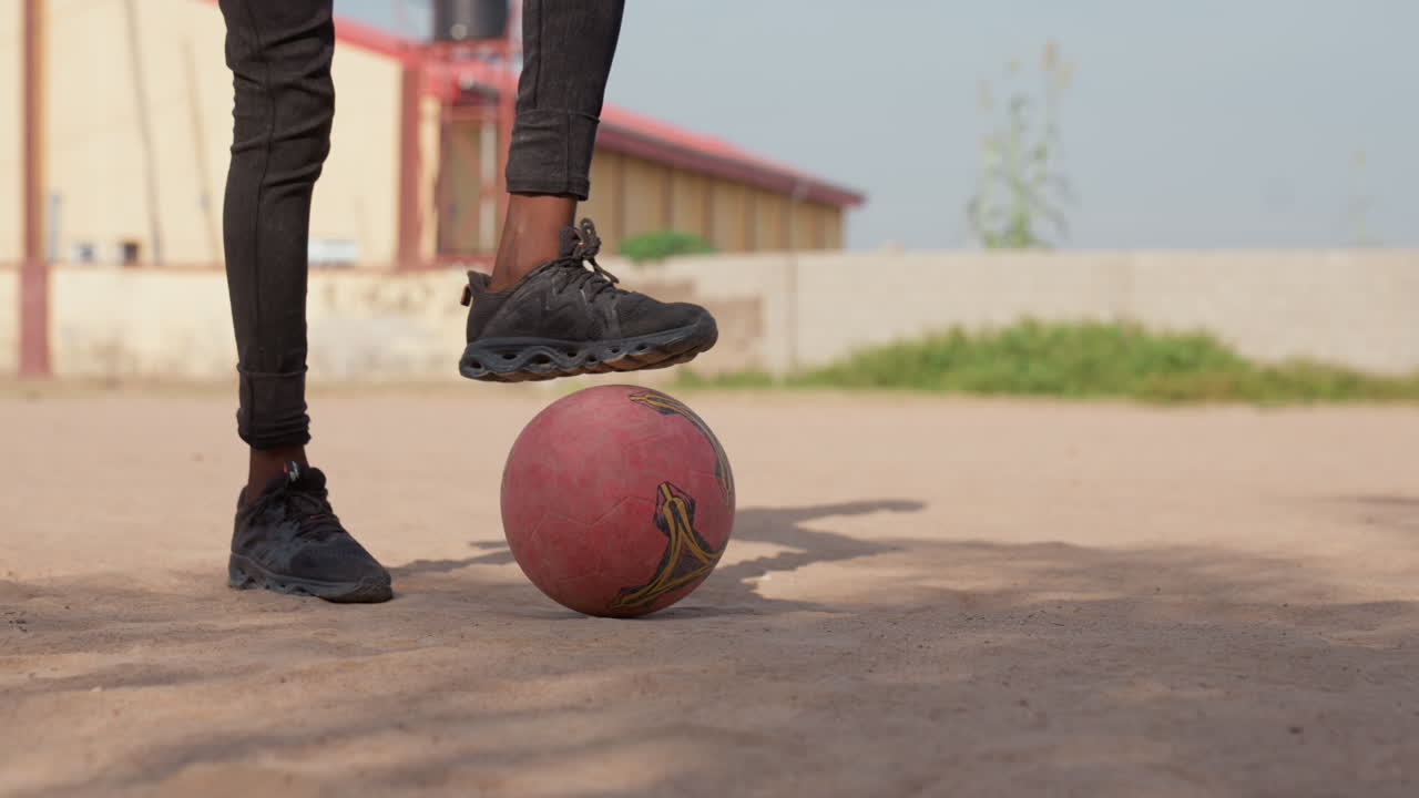 Black youth feet balancing red ball on dusty court, closeup of legs in black sneakers, deliberate footwork and steady control, warm sunlight, school building backdrop, textured dirt surface, focused