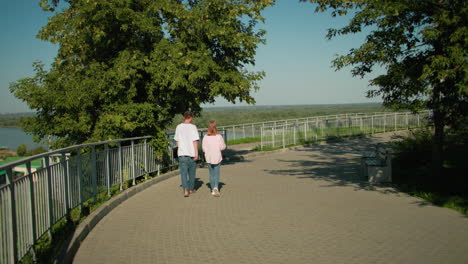 girl in pink top and jeans walking closely with friend holding laptop in left hand along iron rail with scenic background featuring vast forest, flowing river, and clear sky