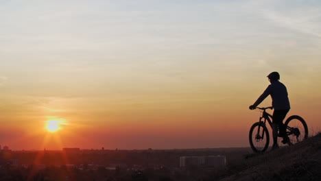 A-male-mountain-biker,-pulls-up-to-a-wood-ramp-atop-a-large-hill-to-observe-a-town-skyline,-as-the-low-sun-silhouettes-his-form