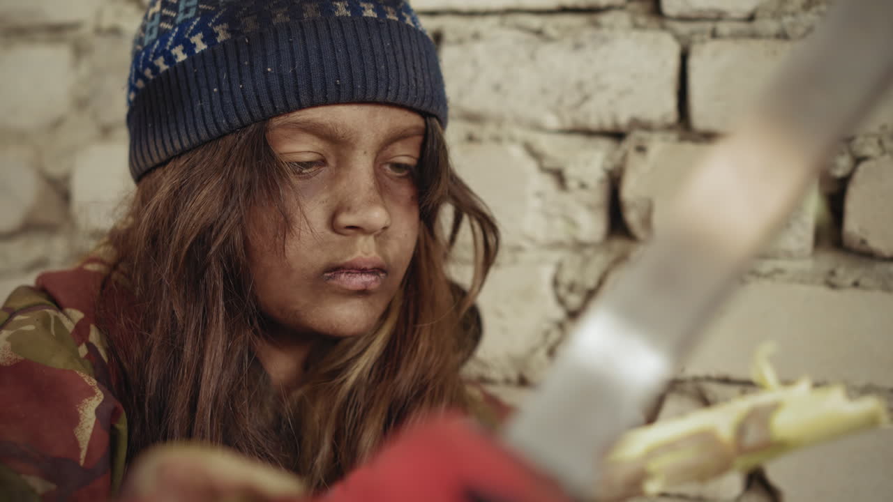 primer plano de un cazador con la cara sucia y gorro de lana, que da forma a un palo áspero con un cuchillo, sentado contra una pared de ladrillo agrietada, con expresión seria y concentrada que revela su instinto de supervivencia