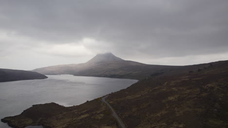 Aerial-View-by-drone-of-mountain-Stac-Pollaidh-with-clouds-above-it-and-Loch-Lurgainn-and-road-along-the-loch-in-Scottish-Highlands