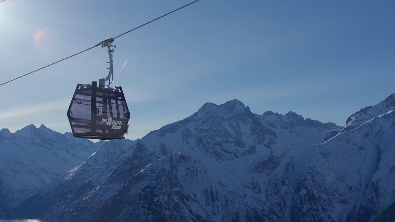 Telecamera che segue l'ascesa di una funivia vuota verso una pista da sci sulla cima di montagne innevate in una giornata di sole.