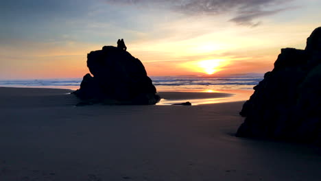 dos personas sentadas sobre una roca alta en la playa de bandon oregon, disfrutando de la puesta de sol.