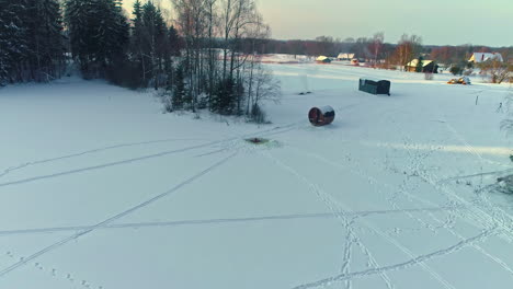 un hombre se sumerge en un agujero cortado en el hielo de un lago congelado por una sauna - revelación aérea de retroceso
