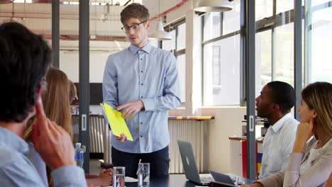 Young-man-holding-file-stands-addressing-team-at-a-meeting