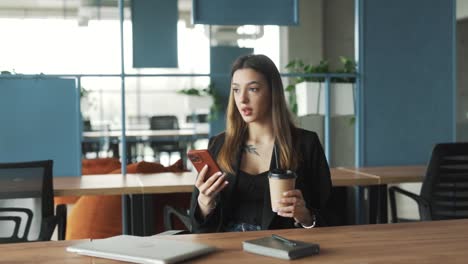 Eine-Junge,-Attraktive-Frau-In-Business-Kleidung-Sitzt-In-Einem-Stilvollen,-Modernen-Coworking-Büro,-Verschickt-Nachrichten-In-Sozialen-Medien-Und-Auf-Dating-Sites-Auf-Ihrem-Smartphone-Und-Nippt-An-Einem-Kaffee