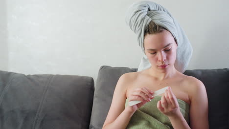 close view of young woman sitting indoors wrapped in green towel with towel on head, calmly filing nails while showing natural skin with visible facial pimples, captured in soft daylight