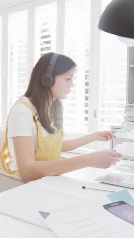 vertical video of biracial woman in headphones using laptop and working at home, slow motion