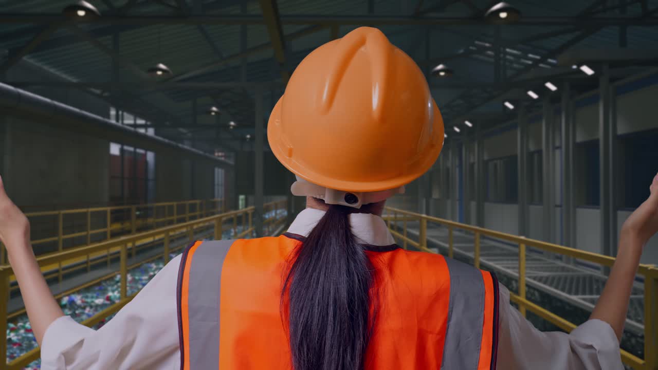 Vista posteriore ravvicinata di un'ingegnere donna con elmetto di sicurezza che allarga le braccia in piedi, mentre ispeziona una fabbrica di riciclaggio della plastica con nastro trasportatore di bottiglie
