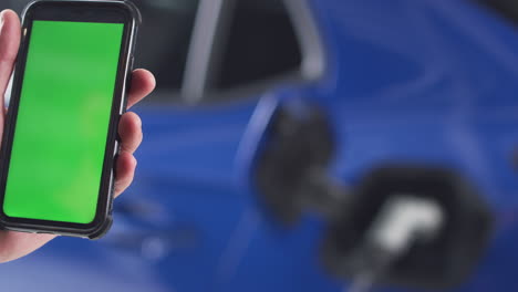 hombre cargando un coche eléctrico con un cable usando un teléfono de pantalla verde para monitorear el nivel de la batería