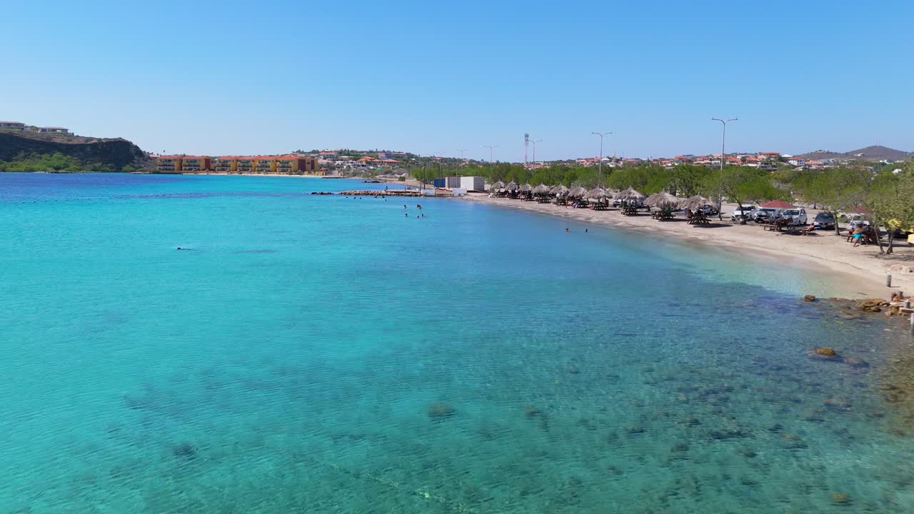 Pristine shoreline at Caracasbaai Beach, Curacao, turquoise waters and lush coastal vegetation under a clear sky with tropical picnic tables on sand