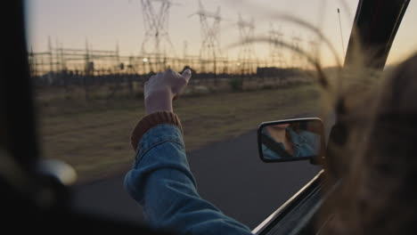 Mujer-Joven-En-El-Coche-Sosteniendo-La-Mano-Por-La-Ventana-Sintiendo-El-Viento-Soplando-Entre-Los-Dedos-Conduciendo-En-El-Campo-En-Un-Viaje-Por-Carretera-Viajando-Para-Vacaciones-De-Verano-Disfrutando-De-La-Libertad-En-La-Carretera-Al-Atardecer