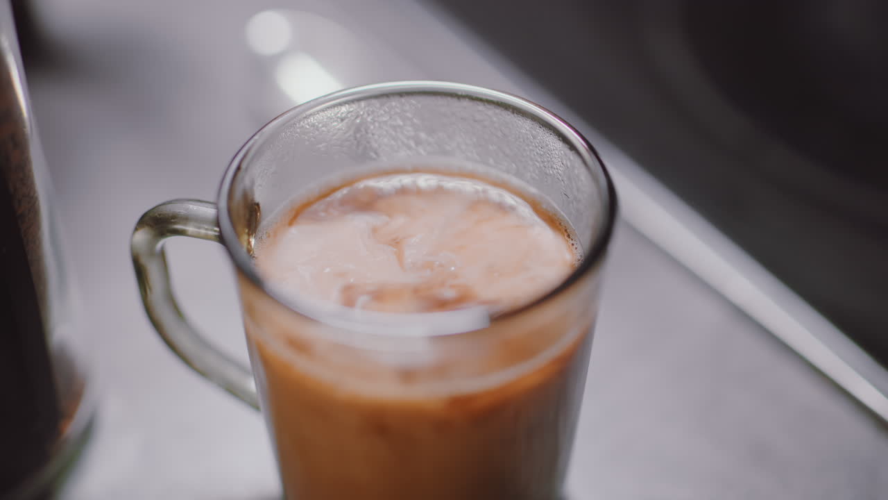 Primo piano di latte cremoso che si versa in un caffè nero bollente dentro una tazza trasparente, il vapore che appanna il vetro, catturando il movimento vorticoso e la fusione di tonalità contrastanti