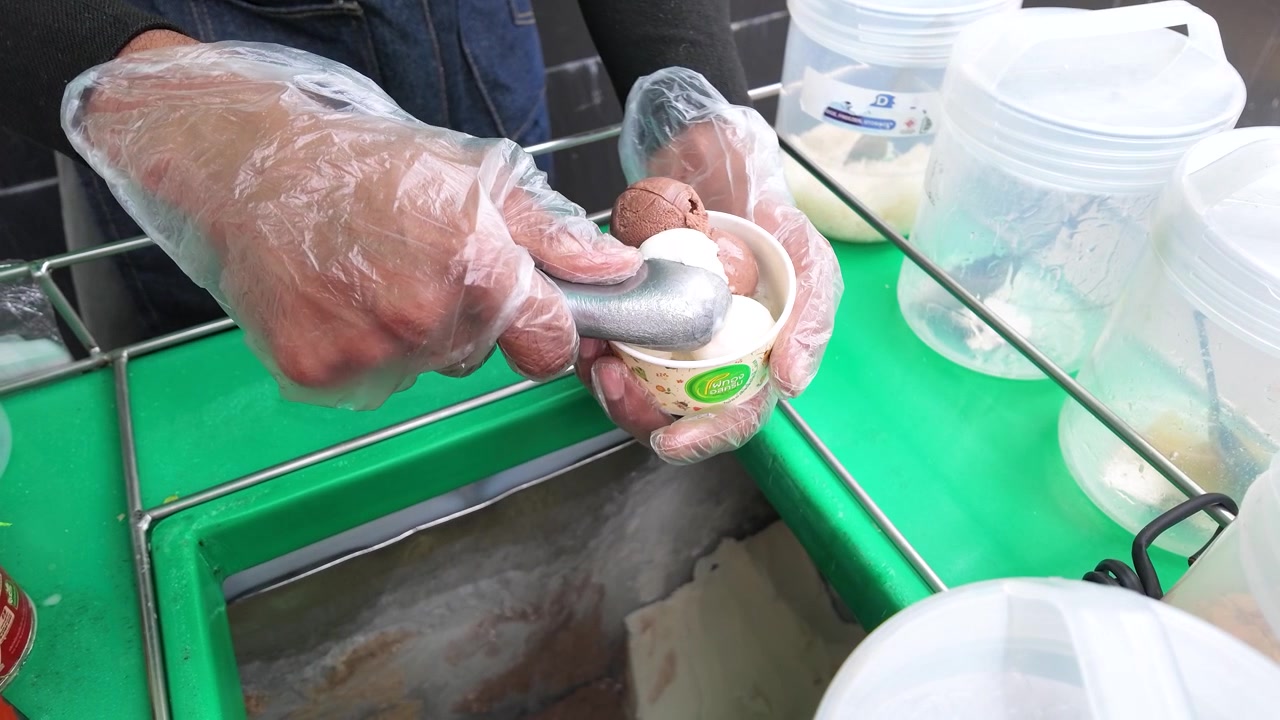 A gloved vendor scoops chocolate and vanilla ice cream into a paper cup in Bangkok.