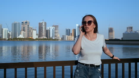 happy beautiful woman in sunglasses and white t-shirt talking on the phone standing on the promenade in the summer against the city and buildings