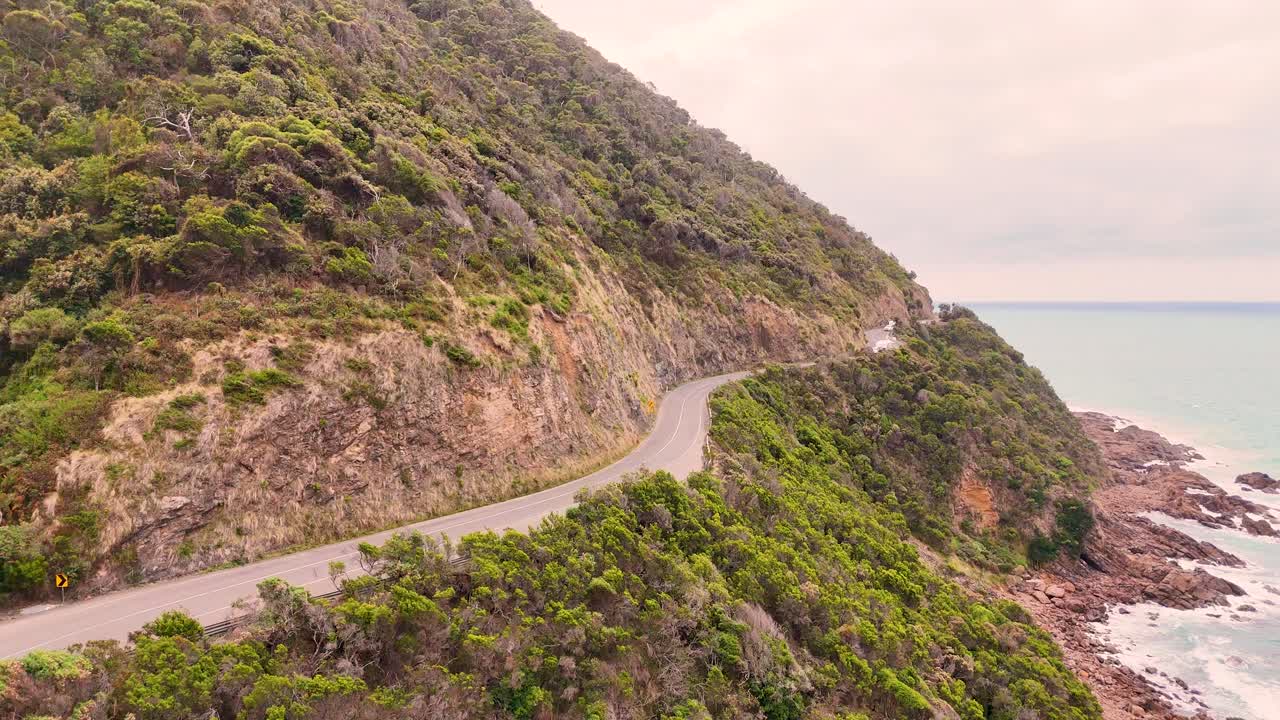 Aerial footage captures the winding Great Ocean Road along Lorne's coastline, showcasing lush greenery and rugged cliffs under soft daylight