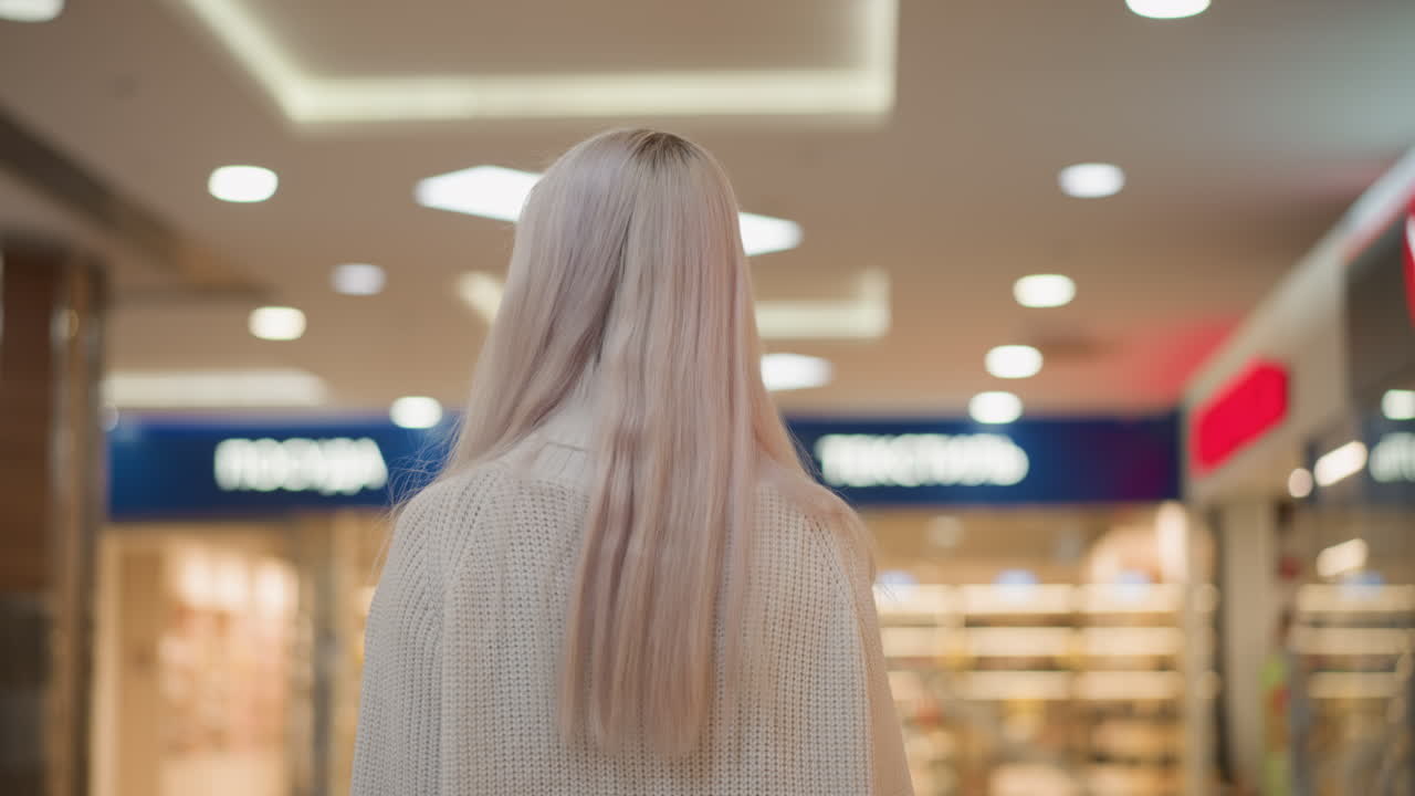 medium back shot of elegant woman with pastel hair walking as she turns head sideways admiring crystal decor and glass railing under bright lights on reflective tiled floor in modern mall corridor