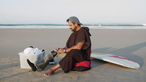 long shot of a male surfer sitting on ocean coast and removing prosthetic leg