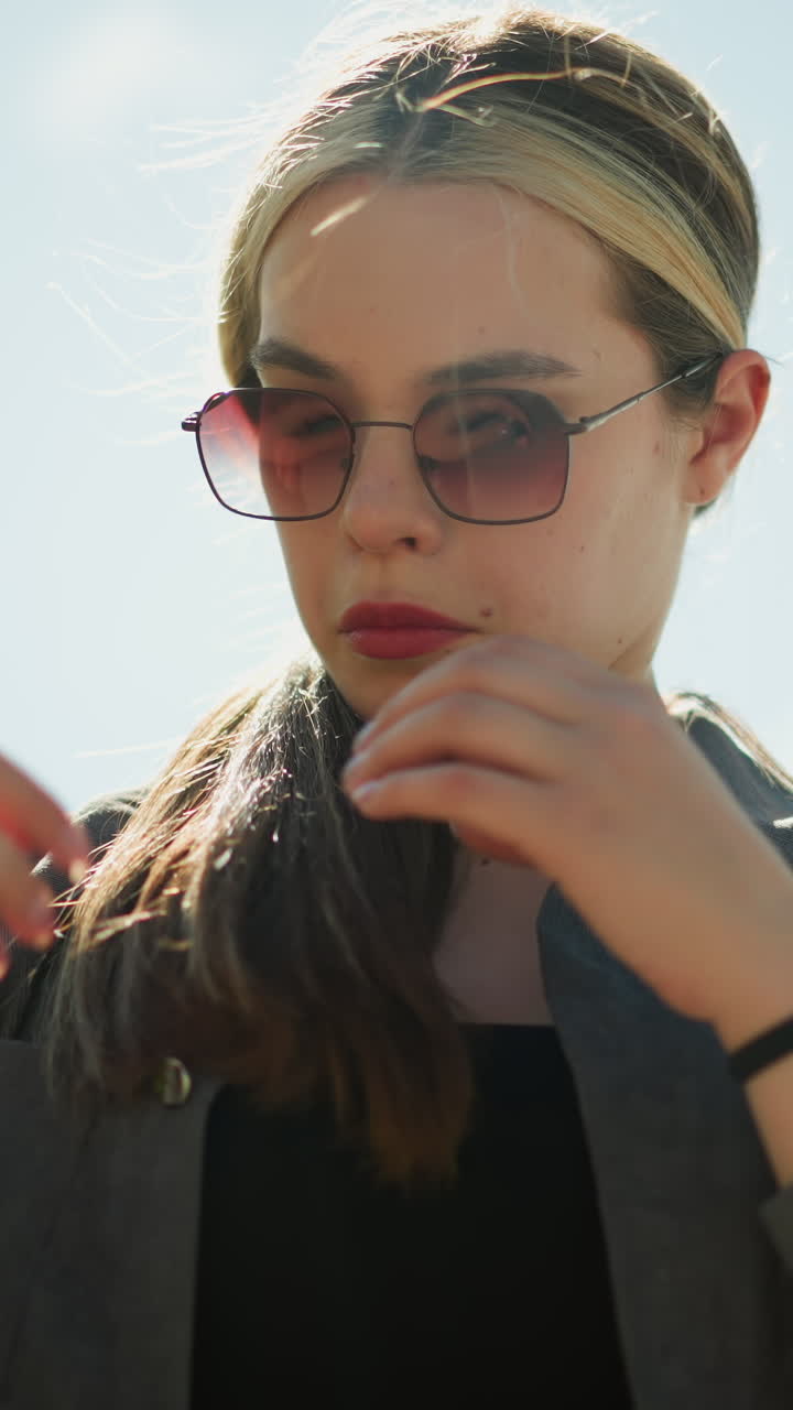 дама в черном кроп-топе под серой рубашкой осторожно держит волосы, отворачивая взгляд, она стоит на улице в солнечный день, ее волосы слегка дрожат на ветру