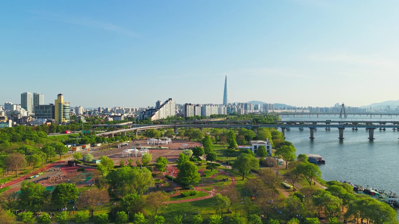 Una ripresa aerea sopra l'Hangang Drone Park rivela le piste da jogging e le aree ricreative lungo il fiume del Parco Gwangnaru Hangang, con la Lotte World Tower nello skyline primaverile di Seul, Corea.