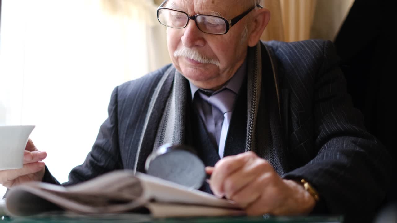 Gray-haired grandfather is sitting in a cafe drinking tea, he is reading a newspaper.