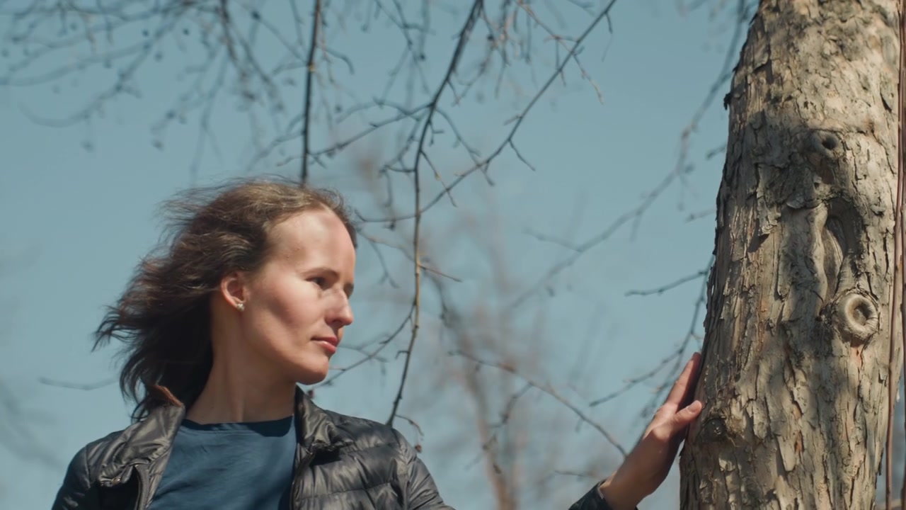 Nahaufnahme einer Frau, die an einem klaren Tag im Freien die raue Baumrinde berührt. Ihr langes Haar bewegt sich leicht im Wind. Sie trägt eine dunkle Jacke und ein blaues Hemd. Im Hintergrund sind kahle Äste vor dem Himmel zu sehen.