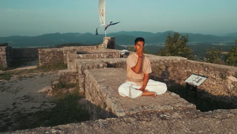 Disparo-De-Un-Dron-De-Un-Hombre-Indio-Sentado-En-Una-Pose-De-Hatha-Yoga-Y-Meditando-Nadhi-Shuddhi-En-La-Pared-Del-Castillo-De-Piedra-En-La-Cima-De-La-Colina-Rodeada-De-Colinas,-Campos-Y-Bosques-En-La-Mañana-Al-Amanecer