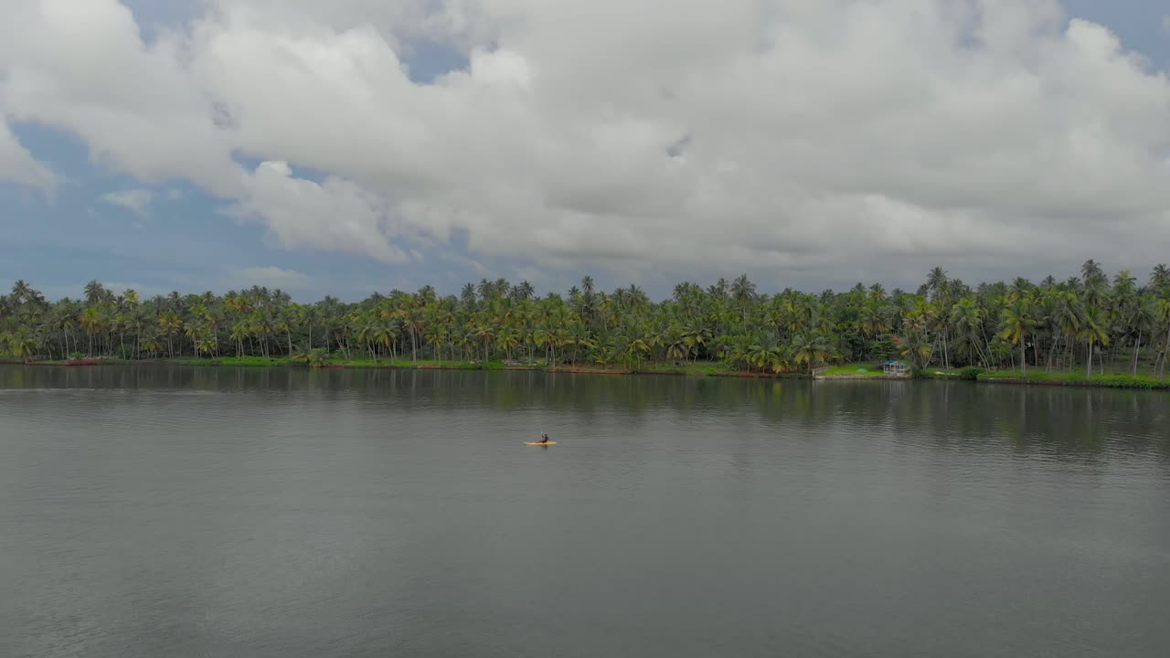 toma aérea de 4k de un hombre indio de 28 años remando en un kayak con equipo de seguridad en los remansos de varkala rodeado de árboles de coco, kerala