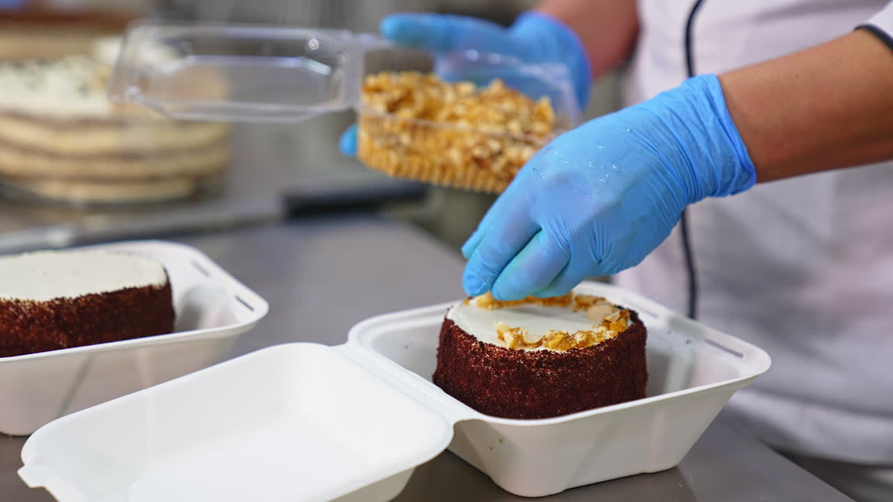 Chocolate cake in individual box being decorated. Gloved hand puts peanuts on top of the dessert. Blurred backdrop.