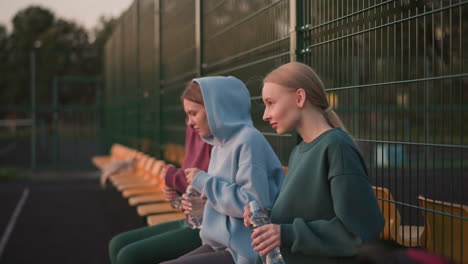 three friends relaxing after exercise, seated with water bottles in their hands as they open them, they are resting after an intense workout session, enjoying a break and hydration