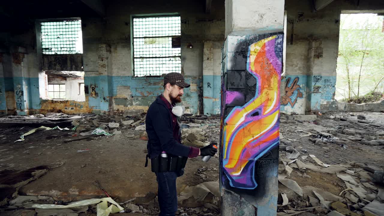 pan tiro de edificio abandonado dañado con altas columnas en el interior y artista de graffiti masculino dibujando imágenes abstractas en el pilar. el hombre tiene guantes de protección y máscara de gas.