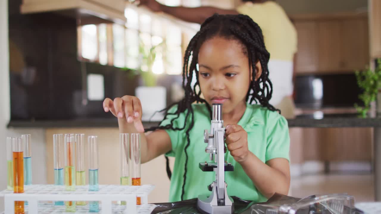 hija afroamericana enfocada usando el microscopio, haciendo experimentos científicos en casa