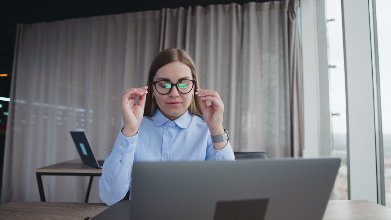 Female employee working on her laptop in the office. Lady takes off glasses, closes laptop and pushes it away looking in the window.